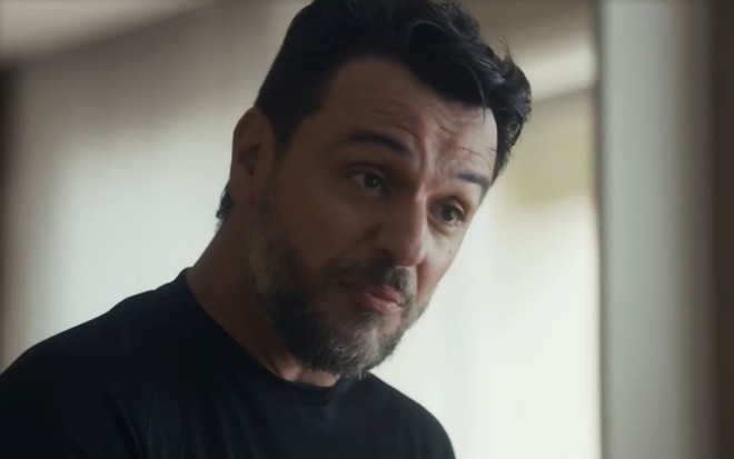 Rodrigo Lombardi usa uma camiseta preta em cena de Travessia; ele dá um sorriso irônico e encara um ponto para além de câmera