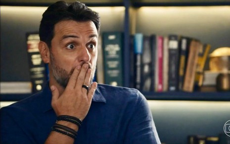 O ator Rodrigo Lombardi está com dedos na boca, expressão de surpresa e usa camisa azul como Moretti em cena de Travessia