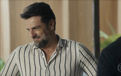 O ator Rodrigo Lombardi está com uma camisa listrada e dá um sorriso debochado em cena de Travessia como Moretti