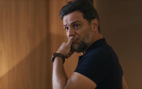 O ator Rodrigo Lombardi estão com a mão na altura da boca e olha desconfiado para alguém fora da imagem em cena de Travessia