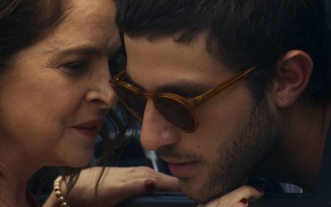 Drica Moraes sussurra no ouvido de Chay Suede; ator usa uma camisa social branca e óculos escuros em cena de Travessia