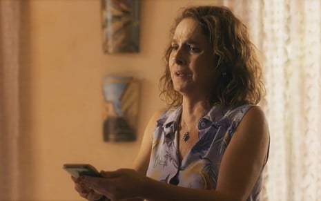 Drica Moraes, caracterizada como Núbia, dedilha um celular em cena de Travessia; ela está empolgada enquanto encara a tela
