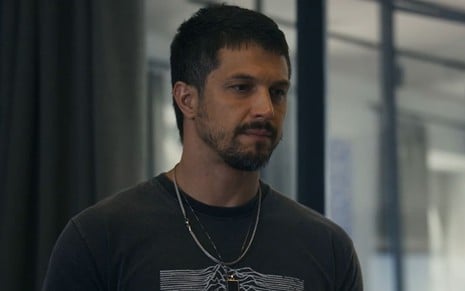 Romulo Estrela tem a expressão séria em cena de Travessia; ele usa uma camiseta preta, justa, e um colar de prata