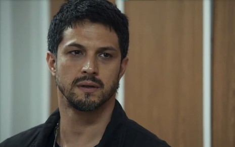 Romulo Estrela caracterizado com Oto em Travessia; ele usa uma camiseta preta e tem o semblante abalado em cena da novela