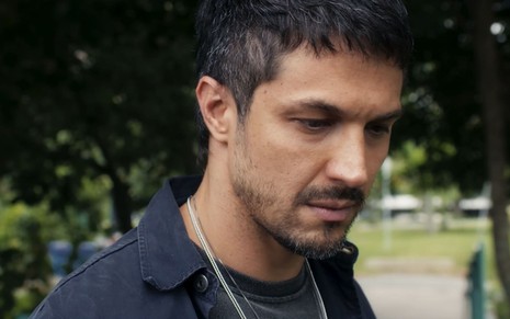 Romulo Estrela usa camiseta e jaqueta pretas e um colar prateado; ele está cabisbaixo em cena de Travessia