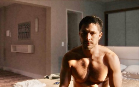 Romulo estrela está sem camisa sentado em uma cama em cena da novela Travessia como seu personagem Oto