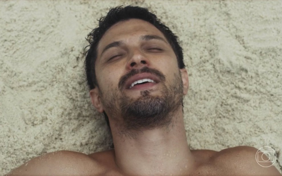 O ator Romulo Estrela está com olhos fechados e boca semiaberta deitado na areia da praia em cena de Travessia como Oto
