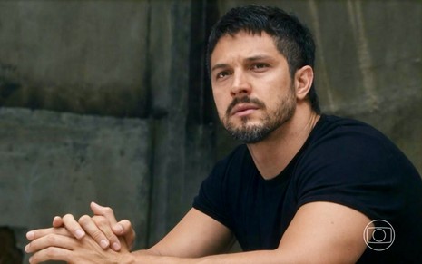 O ator Romulo Estrela está sentado com os braços em cima de uma mesa em cena de Travessia na qual usa camiseta preta justa