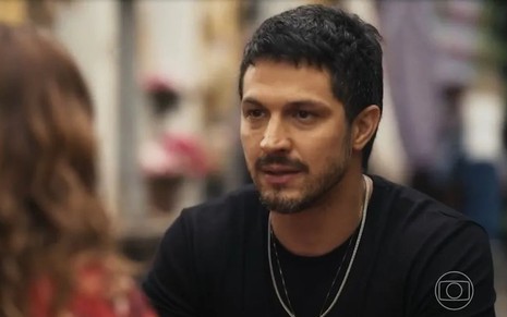 O ator Romulo Estrela usa camiseta preta e está com expressão séria em cena de Travessia como Oto