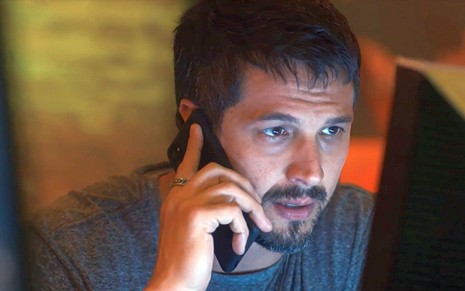 Oto (Romulo Estrela) fala ao telefone diante de um monitor de computador em cena de Travessia