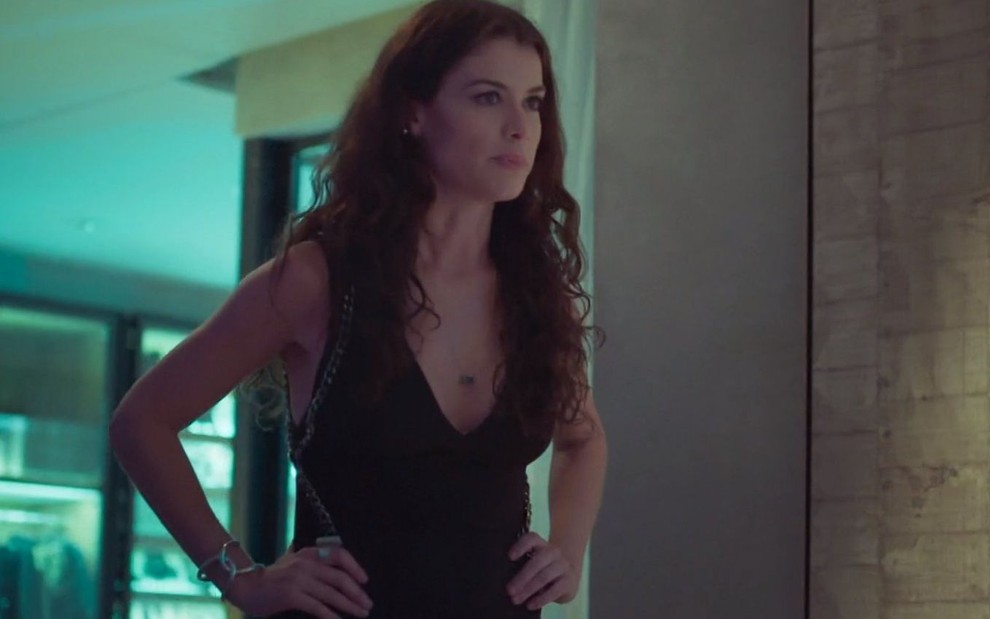 Bárbara (Alinne Moraes) está em pé e com as mãos na cintura em suíte; ela usa um vestido preto em cena de Um Lugar ao Sol