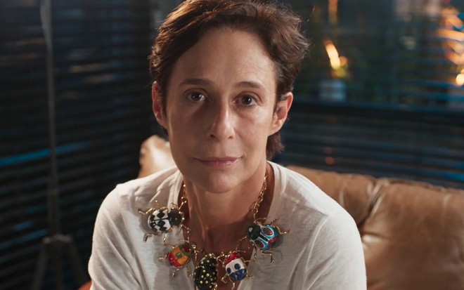 Andrea Beltrão, caracterizada como Rebeca, está sem maquiagem enquanto encara câmera em cena de Um Lugar ao Sol; atriz usa colar de besouros