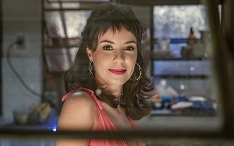 A atriz Andréia Horta está atrás de uma janela basculante que dá para uma cozinha e sorri para a câmera como a Lara em cena de Um Lugar ao Sol