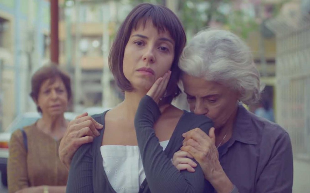 A atriz Andréia Horta com cabelos curtos, chanel, leva a mão direita ao rosto em sinal de desolação enquanto Marieta Severo, a Noca, beija o seu ombro para consolá-la em cena de Um Lugar ao Sol
