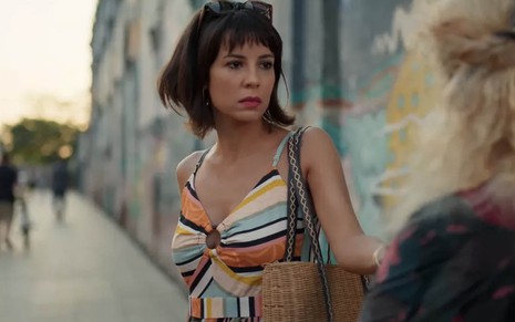 A atriz Andréia Horta caracterizada como Lara, no meio de uma rua, com um muro pichado ao fundo e expressão de surpresa em cena de Um Lugar ao Sol
