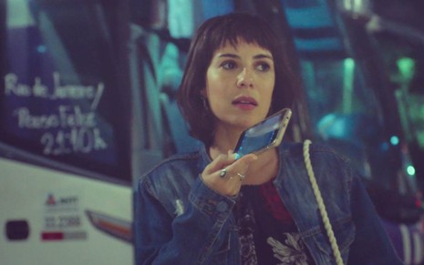 A atriz Andréia Horta como Lara segura um celular com a mão direita e envia uma mensagem de voz com um ônibus de viagem ao fundo em uma rodoviária em cena de Um Lugar ao Sol