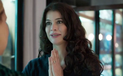 Alinne Moraes em cena de Um Lugar ao Sol: atriz está bata preta, cabelo solta, sorri e junta as mãos em sinal de agradecimento