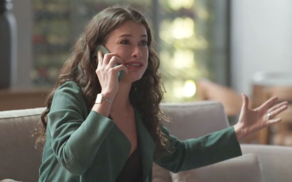 Alinne Moraes em cena de Um Lugar ao Sol: atriz está com camisa verde, cabelo solto e fala ao telefone com expressão de desespero