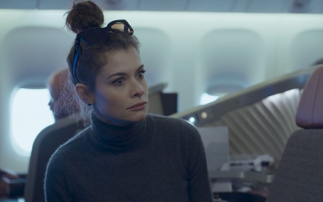 Alinne Moraes grava com cabelo preso, blusa cinza de gola alta e expressão de surpresa como Bárbara de Um Lugar ao Sol