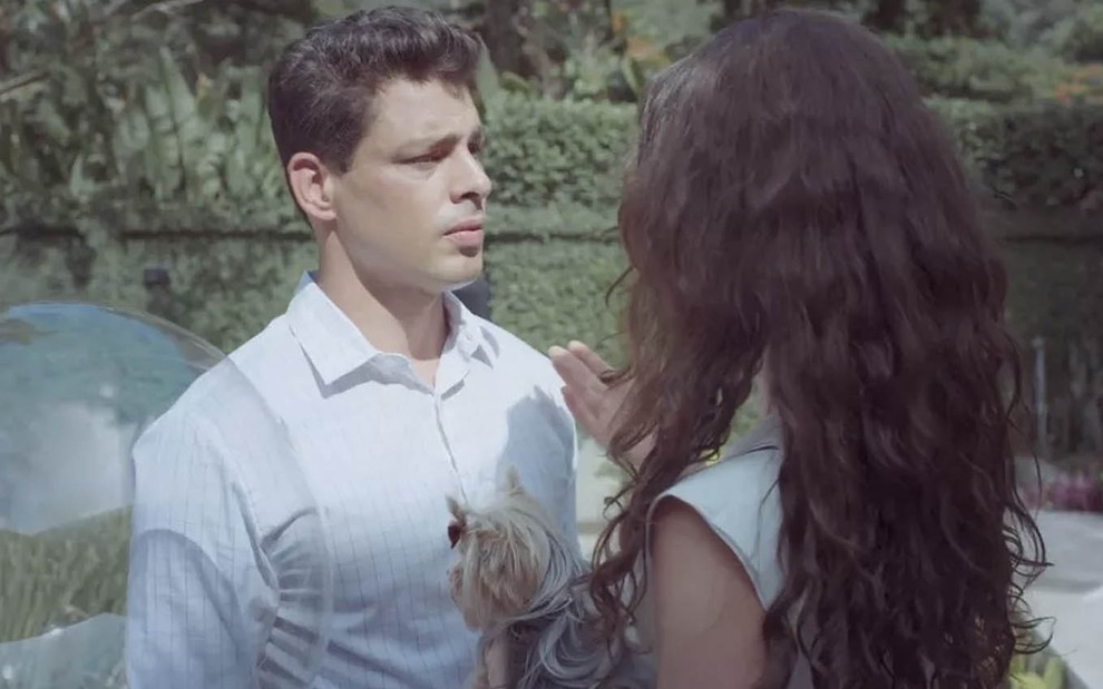 O ator Cauã Reymond com Christian em um jardim enquanto fala com Alinne Moraes, a Bárbara, de costas, segurando um cachorro no colo em cena de Um Lugar ao Sol