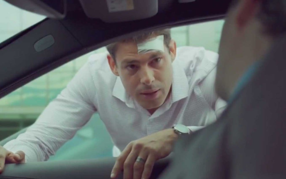 O ator Cauã Reymond caracterizado como Christian se apoia na janela de um carro com um curativo na testa em cena de Um Lugar ao Sol