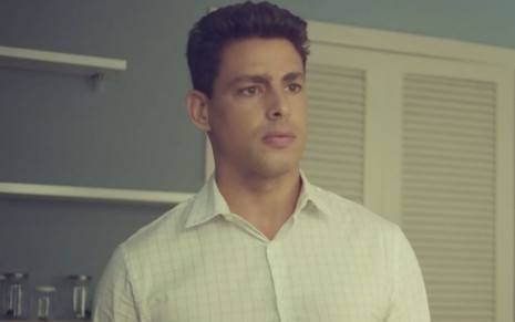 Cauã Reymond em cena de Um Lugar ao Sol: ator está caracterizado como Renato/Christian, camisa social branca e olha para alguém fora do quadro