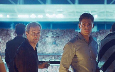 Túlio (Daniel Dantas) e Christian (Cauã Reymond) estão em camarote em estádio de futebol em cena de Um Lugar ao Sol