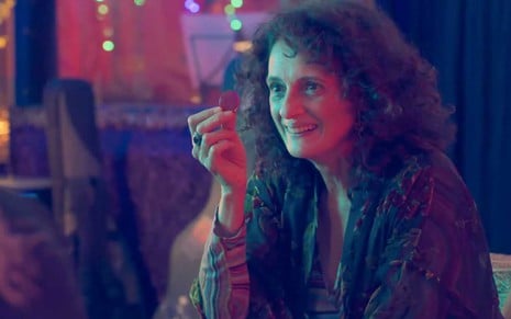 A atriz Denise Fraga segura uma moeda com a mão direita e a exibe com um sorriso no rosto como Júlia em cena de Um Lugar ao Sol