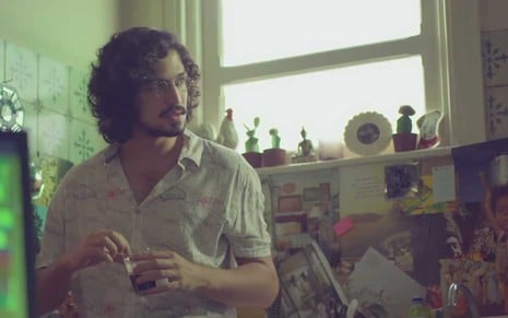 O ator Gabriel Leone está em uma cozinha, apoiado em uma pia, com os cabelos soltos, na altura do pescoço, cacheados e pretos, com óculos redondos como Felipe em cena de Um Lugar ao Sol