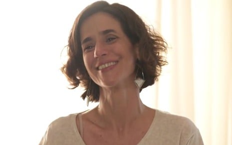 Mariana Lima em cena de Um Lugar ao Sol: caracterizada com camisa cinza, atriz sorri para alguém fora do quadro