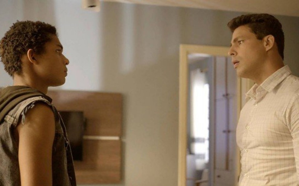 Ravi (Juan Paiva) e Christian (Cauã Reymond) estão em pé na sala do apartamento do motorista em cena de Um Lugar ao Sol