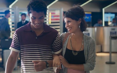 Ravi (Juan Paiva) e Lara (Andréia Horta) de braços dados sorrindo no saguão do aertoporto