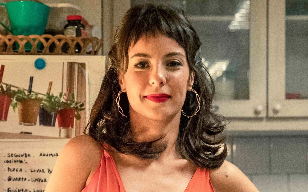 Andréia Horta caracterizada como Lara em Um Lugar ao Sol: atriz está de pé, usa regata laranja, cabelo curto com franja, brincos de argola e está maquiada com batom vermelho