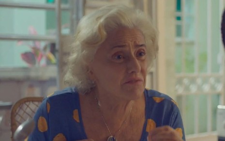 Marieta Severo caracterizada como Noca em Um Lugar ao Sol: atriz está com camisa azul com detalhes em amarelo e olha indignada para alguém fora do quadro