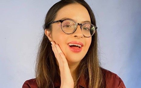 Iluminada por luz quente, Samantha Quadrado sorri e apoia uma das mãos na bochecha; ela usa óculos com aro preto e blusa vermelha