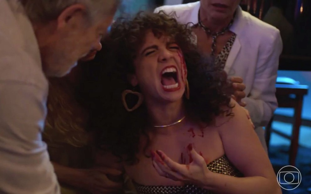 Renata Gaspar como Stephany em cena de Um Lugar ao Sol: atriz está em close, com cabelos cacheados, batom vermelho e grita com parte do rosto ensangüentado