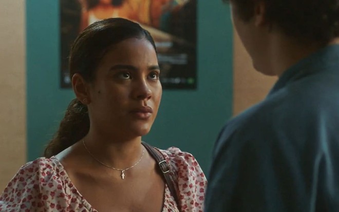 Bella Campos usa uma blusa florida com mangas curtas e bufantes; ela tem o cabelo amarrado num rabo de cavalo baixo e ostenta um semblante sério em cena de Vai na Fé