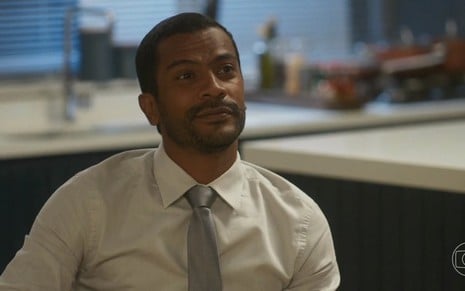 O ator Samuel de Assis está de camisa branca com gravata cinza e olha feliz para a frente em cena da novela Vai na Fé