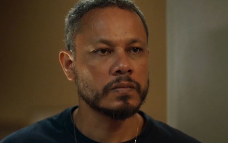 Che Moais como Carlão em Vai na Fé; ele usa uma camiseta polo preta e tem o semblante sério em cena da novela