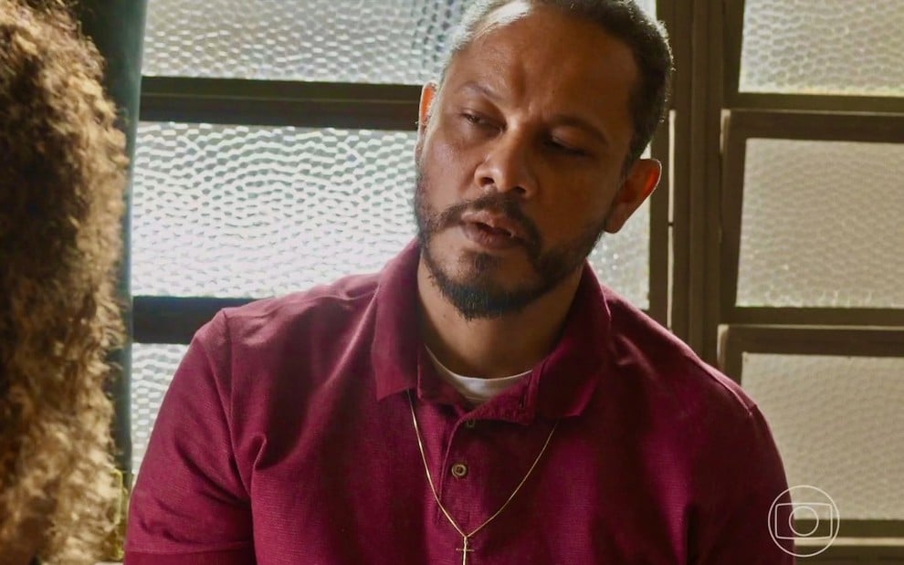 O ator Che Moais está usando uma camisa polo beterraba e faz uma expressão séria em cena como Carlão em Vai na Fé
