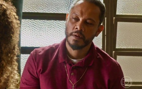 O ator Che Moais está usando uma camisa polo beterraba e faz uma expressão séria em cena como Carlão em Vai na Fé