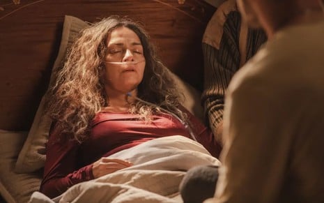 Claudia Ohana caracterizada como Dora em cena de Vai na Fé; ela está deitada, com uma sonda nasogástrica, os olhos fechados e os os cabelos soltos