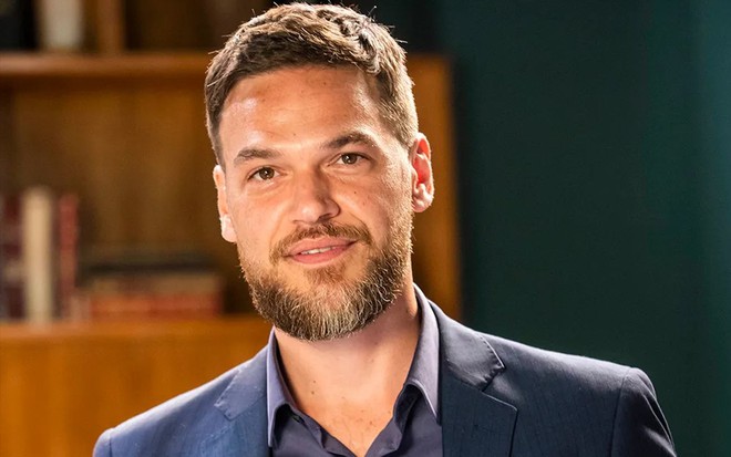 Com paletó e camisa social, Emilio Dantas tem expressão neutra no cenário de Vai na Fé