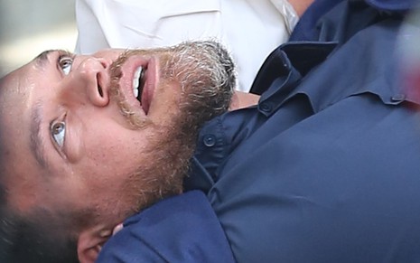 Emilio Dantas está caído com olhos arregalados e boca entreaberta durante gravação de cena da novela Vai na Fé