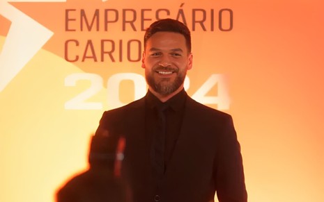Emilio Dantas caracterizado como Theo em Vai na Fé; ele sorri na frente de um banner com o termo "Empresário carioca do ano 2024"