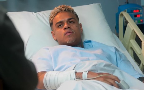 MC Cabelinho caracterizado como Hugo; ele está abatido e deitado numa maca de hospital em cena de Vai na Fé