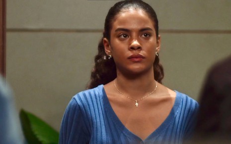 A atriz Bella Campos está com uma camiseta azul e olha com cara de séria para a frente em cena da novela Vai na Fé