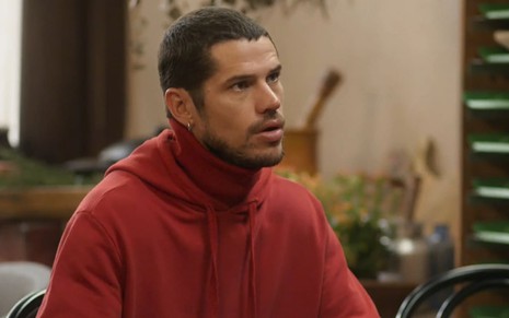 José Loreto caracterizado como Lui Lorenzo em Vai na Fé; ele está com um moletom vermelho e parece sério em cena de Vai na Fé