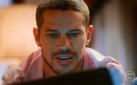 O ator José Loreto olha para a tela de um computador com um leve sorriso em cena da novela Vai na Fé, da Globo