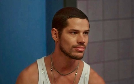 O ator José Loreto usa regata branca e olha para alguém fora da imagem em cena da novela Vai na Fé